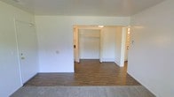 This single-floor space offers a straightforward and functional layout designed for comfortable everyday living. The main living area features smooth hardwood-style flooring that transitions seamlessly from a carpeted entry zone, creating a clear visual distinction between spaces. Bright white walls throughout lend a clean, neutral backdrop that enhances the sense of openness and allows natural light to reflect throughout the interior.

A central opening connects the main living area to an inner hallway or secondary room, where a built-in closet with double doors provides convenient storage. The closet's open design reveals a practical shelving rod setup, ideal for organized storage. Beyond the closet, the hallway extends further, hinting at additional rooms — glimpses of what appear to be a kitchen and another area are visible in the background, suggesting a well-connected floor plan.

The ceiling is smooth and unadorned, maintaining a consistent height that contributes to the overall airy feel of the space. Baseboard heating units are visible along the walls, indicating a reliable in-unit heating system. The minimalist design of the space, with its clean lines and neutral palette, provides a versatile foundation that can be easily personalized to suit a variety of interior styles and needs.