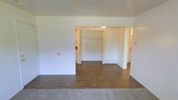 This single-floor space offers a straightforward and functional layout designed for comfortable everyday living. The main living area features smooth hardwood-style flooring that transitions seamlessly from a carpeted entry zone, creating a clear visual distinction between spaces. Bright white walls throughout lend a clean, neutral backdrop that enhances the sense of openness and allows natural light to reflect throughout the interior.

A central opening connects the main living area to an inner hallway or secondary room, where a built-in closet with double doors provides convenient storage. The closet's open design reveals a practical shelving rod setup, ideal for organized storage. Beyond the closet, the hallway extends further, hinting at additional rooms — glimpses of what appear to be a kitchen and another area are visible in the background, suggesting a well-connected floor plan.

The ceiling is smooth and unadorned, maintaining a consistent height that contributes to the overall airy feel of the space. Baseboard heating units are visible along the walls, indicating a reliable in-unit heating system. The minimalist design of the space, with its clean lines and neutral palette, provides a versatile foundation that can be easily personalized to suit a variety of interior styles and needs.