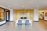 Located at 3112 Leeland St in Houston, TX, this single-floor commercial space serves as the lobby and entry hub for Coopeado. The layout encompasses a hallway and several versatile rooms, all unified by a clean, contemporary design aesthetic.

The reception area immediately establishes the space's identity, featuring a sleek white desk paired with upholstered gray chairs on both sides. A striking textured wood-panel accent wall with vertical gold trim serves as the backdrop, prominently displaying the Coopeado logo. Recessed lighting illuminates the area from above, while large-format neutral floor tiles extend throughout the entire space.

The hallway transitions seamlessly from the reception area, where a decorative wall arrangement of geometric, diamond-shaped planters adds a natural, artistic element against crisp white walls. Dark navy blue baseboard accents provide visual contrast throughout.

Floor-to-ceiling glass walls and oversized windows flood the space with natural light and offer open views of the surrounding outdoor landscape, including a mature tree visible from the main corridor. A seating area near the elevator features a black leather chair, potted indoor plants, and framed wall art, creating a welcoming waiting space. The overall ambiance is polished and professional, balancing modern minimalism with warm, thoughtful design details.