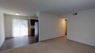 This single-floor residence offers a practical and functional layout designed for comfortable everyday living. The main living area is finished with neutral wall-to-wall carpeting, creating a warm and understated atmosphere. A partial dividing wall separates the living space from the kitchen, maintaining an open feel while providing a sense of definition between the two areas. The kitchen features dark wood cabinetry, a stainless steel refrigerator, and hard-surface flooring, offering a clean contrast to the carpeted living room. A large window with vertical blinds allows natural light to filter into the kitchen and adjacent living space, brightening the interior. A hallway, visible from the main living area, leads to the additional rooms of the unit, suggesting a thoughtfully segmented floor plan. Overhead lighting in the hallway and a visible ceiling vent indicate practical utility throughout. The overall ambiance is clean, neutral, and move-in ready, with a straightforward design that provides a solid foundation for personalization and furnishing.