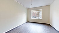 This well-organized single-floor unit offers a practical and thoughtfully laid out living space. The layout is anchored by a generously sized bedroom, which accounts for the majority of the floor area and features large windows that frame views of the surrounding neighborhood streetscape, allowing natural light to fill the room. The walls are finished in soft, neutral tones with clean white trim and baseboards, complementing the dark hardwood-style flooring that runs consistently throughout the space.

A narrow but functional hallway connects the key areas of the unit, featuring built-in open shelving niches in the walls — ideal for storage or display. The hallway is equipped with a ceiling light fixture and leads to both the bedroom and the bathroom. A coat hook panel is also visible near the bedroom entry, adding practical storage options.

The bathroom showcases a classic design with white subway tile surrounding a full soaking bathtub and shower combination. The floor features a charming mosaic tile pattern in soft green and white tones, adding a vintage character to the space. A recessed wall niche provides additional storage within the shower area.

The unit also includes an entry hallway with warm-toned flooring and access to built-in shelving, completing a compact but complete residential layout designed for efficient, comfortable living.