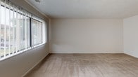 This single-story residence offers a straightforward, functional layout across several distinct areas. The living area is generously proportioned, finished with wall-to-wall beige carpet and neutral cream-toned walls that create a clean, understated backdrop. A large window fitted with vertical blinds allows natural light to filter into the space, offering views of the surrounding exterior. An adjacent carpeted room, similarly finished with neutral walls and a window with vertical blinds, provides a bright and open atmosphere suitable for use as a bedroom.

A second carpeted room is accessible via a short hallway and features a built-in closet, adding practical storage to the space. The bathroom is lined with classic white subway tile on the lower walls and floors finished with a gray-and-white mosaic tile pattern, lending a timeless, clean aesthetic. A vanity with storage is situated along one wall.

The kitchen is equipped with dark wood cabinetry offering ample storage, light-toned countertops, a stainless steel single-basin sink, and under-cabinet lighting. A dishwasher is integrated into the cabinetry. The overall ambiance of the unit is neutral and move-in ready, with consistent finishes throughout that create a cohesive, functional living environment.