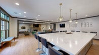 Located at Grove Park in Columbus, Georgia, this single-floor community space offers a generously sized, open-concept layout combining a fully equipped kitchen and a welcoming living room. The kitchen is a standout feature, anchored by an expansive marble-top island that comfortably seats multiple guests on sleek black bar stools. Star-shaped pendant lights hang above, casting warm, decorative light over the space. The kitchen includes stainless steel appliances—a full-size refrigerator, oven, and microwave—set against crisp white cabinetry and teal subway tile backsplash. Dark crown molding provides a refined contrast to the light gray walls and warm hardwood-style flooring throughout. The living area is equally impressive, centered around a striking floor-to-ceiling stone fireplace. Comfortable seating, including a gray sofa, green accent chair, and a patterned area rug, creates an inviting atmosphere. A round dining table with black chairs and a wall-mounted TV add to the room's versatility. Architectural columns with dark paneling add visual interest, while a wall of glass-paned French doors floods the space with natural light and offers views of the surrounding greenery.