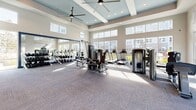 This single-floor fitness center is thoughtfully designed to support a full range of exercise activities in a bright, welcoming environment. The main gym area is expansive, featuring soaring coffered ceilings with large ceiling fans and an array of natural light streaming in through floor-to-ceiling windows that frame views of the surrounding outdoor spaces. The gym is well-equipped with a comprehensive selection of strength-training machines, free weights with benches, and cardio equipment including treadmills and ellipticals from brands such as TRUE Fitness. The flooring is a durable, patterned carpet suited for high-traffic fitness use. A separate studio or yoga room adjoins the main floor, enclosed by full-height glass walls that maintain visual openness while providing a defined, versatile space for group classes or individual practice. This studio features smooth hardwood flooring, roller shades on its large sliding glass doors, and a tray ceiling with recessed lighting. Back in the main area, wall-mounted flat-screen televisions are positioned for easy viewing during workouts. The space is flooded with natural sunlight throughout the day, contributing to an energizing and motivating atmosphere. Direct access to an outdoor patio or pool area is available through glass doors, seamlessly connecting indoor fitness activities with the outdoor amenities.