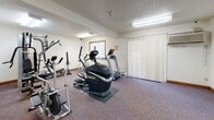 This fitness center at 1821 15th Ave SE in Saint Cloud, Minnesota, offers a well-equipped, single-floor exercise facility designed to support a wide range of workout routines. The space features plush, multi-tone carpeted flooring throughout, warm white walls with wood baseboards, and a textured ceiling fitted with bright fluorescent lighting that keeps the entire room well-illuminated.

The cardio area is stocked with several machines, including ellipticals, a stationary bike, and a treadmill, providing ample options for aerobic exercise. A dedicated strength training section houses a multi-station cable machine along with a rack of dumbbells, kettlebells, resistance bands, and a medicine ball, all neatly organized on a wheeled storage rack. A barbell with weight plates is also available for free weight training.

A mirrored wall with a ballet-style barre adds a functional stretch and balance area, complemented by stability balls for core and flexibility work. An elevated wall-mounted television provides entertainment during workouts, while a wall-mounted air conditioning unit ensures comfortable temperature control year-round.

Practical amenities include a built-in drinking fountain, a hand sanitizer dispenser, and a waste bin, promoting hygiene and convenience. Informational posters and facility rules are displayed on the walls. A window dressed with vertical blinds and a secondary entry door complete this thoughtfully arranged fitness space.