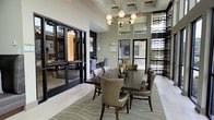 This elegantly designed single-floor space exudes a contemporary yet welcoming atmosphere, thoughtfully curated for both social gatherings and productive work. The centerpiece of the main area is a substantial dark wood dining table surrounded by plush, upholstered leather chairs, all anchored by a textured wave-patterned area rug. Above, a striking mid-century modern chandelier with globe bulbs serves as a refined focal point, complementing the elevated coffered ceiling that adds architectural depth and a sense of grandeur to the room. Large black-framed windows and glass doors flood the interior with natural light while offering views of the surrounding exterior, creating a seamless connection between indoors and out. Sheer, striped drapery panels frame the windows, softening the space with an added layer of texture. Adjacent to the dining area, a dedicated workspace is outfitted with individual workstations and ergonomic seating, making it ideal for focused tasks. The light hardwood flooring runs throughout, tying the spaces together with a clean, modern aesthetic. The overall ambiance is polished and inviting, balancing functional design with upscale finishes.