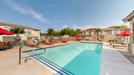 Located at 19955 N 51st Ave in Phoenix, Arizona, this outdoor community pool area offers a well-appointed recreational retreat designed for the Southwest lifestyle. The centerpiece is a generously sized swimming pool with clear turquoise water, tiled edges, and built-in entry steps with safety rails, making it accessible and inviting. The surrounding pool deck is finished with paved brick and concrete surfaces, providing ample space for lounging and movement. Multiple sun loungers and chairs with vibrant cushions are positioned around the pool deck, along with red shade umbrellas for relief from the Arizona sun. A covered patio area adjacent to the pool features recessed lighting, a brick-paved floor, and comfortable seating with colorful striped cushions arranged around a wrought iron coffee table — ideal for shaded relaxation. The entire area is enclosed by a white metal fence with a secure gate, ensuring privacy and safety. Mature palm trees, desert shrubs, and ornamental plantings accent the perimeter, complementing the surrounding Southwest-style architecture characterized by stucco exteriors and terra cotta tile rooflines. The space benefits from abundant natural sunlight and open sky views, embodying the quintessential outdoor living experience of the Phoenix metro area.