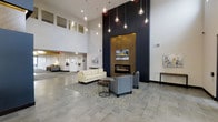 This thoughtfully designed single-floor space makes a striking first impression with its soaring double-height ceilings and expansive, open layout. The centerpiece of the main lounge area is a bold, floor-to-ceiling accent wall finished in deep charcoal, framing a herringbone wood-paneled surround that houses a sleek linear fireplace. A curated arrangement of globe pendant lights cascades from the ceiling at varying heights, casting warm, ambient illumination throughout the space. The seating area features a clean-lined cream sofa and a contemporary gray accent chair, arranged over a textured area rug atop large-format tile flooring in a cool gray tone. A minimalist side table and small round coffee table complete the lounge vignette. The open-plan layout extends beyond the main seating area, revealing additional lounge and activity zones in the background, all bathed in natural light from clerestory windows positioned high on the walls. The overall aesthetic is modern and sophisticated, balancing neutral tones with warm wood accents and polished fixtures to create a welcoming, upscale communal environment suited for relaxing or socializing.