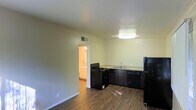 This single-floor space features an open layout that seamlessly connects the kitchen and living area, creating a functional and practical environment. The kitchen is equipped with black appliances, including a gas range, dishwasher, and refrigerator, complemented by dark cabinetry and light-toned countertops. A ceiling-mounted light fixture provides direct illumination over the kitchen workspace, while a second overhead light brightens the adjoining area. Dark wood-style flooring runs throughout, offering a consistent and clean aesthetic. The walls are finished in a neutral light tone, contributing to a bright, airy feel. A doorway visible from the main area leads to an adjacent room with warm lighting, suggesting additional living or utility space beyond. Ceiling-mounted HVAC vents indicate a central air system, adding to the unit's practical features. The open plan layout maximizes the usable space, making it well-suited for everyday living.