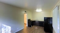 This single-floor space features an open layout that seamlessly connects the kitchen and living area, creating a functional and practical environment. The kitchen is equipped with black appliances, including a gas range, dishwasher, and refrigerator, complemented by dark cabinetry and light-toned countertops. A ceiling-mounted light fixture provides direct illumination over the kitchen workspace, while a second overhead light brightens the adjoining area. Dark wood-style flooring runs throughout, offering a consistent and clean aesthetic. The walls are finished in a neutral light tone, contributing to a bright, airy feel. A doorway visible from the main area leads to an adjacent room with warm lighting, suggesting additional living or utility space beyond. Ceiling-mounted HVAC vents indicate a central air system, adding to the unit's practical features. The open plan layout maximizes the usable space, making it well-suited for everyday living.