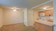 This single-floor space offers an open-concept design that seamlessly connects the kitchen and living area. The living room features wall-to-wall carpeting in a neutral beige tone, providing a warm and comfortable underfoot feel. A ceiling fan with a light fixture serves as the central overhead lighting source, contributing to both airflow and illumination in the space.

The kitchen is positioned adjacent to the living area and is partially separated by a raised breakfast bar counter, offering a natural division between the two spaces while maintaining an open feel. The kitchen is equipped with white appliances, including a range and an over-the-range microwave, and is complemented by warm-toned wooden cabinetry that provides ample storage. The countertops carry a light, neutral finish consistent with the overall color palette of the space.

A structural column and partial wall near the center of the layout define the transition zones within the unit. Visible HVAC vents are integrated into the walls, indicating a central climate control system. The overall ambiance is clean, functional, and neutral, making it a versatile and move-in-ready space.