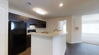 This single-floor residence offers a clean, functional layout centered around an open-concept design that seamlessly connects the kitchen and living areas. The kitchen is a standout feature, showcasing rich dark espresso cabinetry that contrasts beautifully with the light-toned countertops along the peninsula breakfast bar. The peninsula provides both additional prep space and a casual dining or seating area, making it a natural gathering point. A built-in sink with a brushed bronze faucet is integrated into the peninsula, adding convenience and a touch of style. Black appliances, including a refrigerator and range, complement the dark cabinetry for a cohesive aesthetic. Recessed lighting and a flush-mount ceiling fixture ensure the kitchen is well-lit throughout. Dark hardwood-style flooring flows continuously through the main living areas, lending warmth and visual continuity to the space. The living area benefits from a window that invites natural light, contributing to an open and airy feel. A hallway visible from the kitchen leads to additional rooms, hinting at a practical and well-organized floor plan. Neutral wall tones throughout keep the interior feeling bright and versatile, ready to complement a variety of furnishing styles.