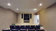 This single-floor space is designed as a dedicated screening or presentation room, offering a focused and functional environment suited for group viewings or presentations. The room is anchored by a large flat-screen display mounted within a built-in media wall, flanked by floor-standing surround sound speakers that deliver an immersive audio-visual experience. Below the display, a built-in console provides space for media equipment, adding to the room's organized and purposeful layout. Rows of dark upholstered chairs are arranged in a theater-style configuration, providing clear sightlines to the display from every seat. The walls are finished in a warm neutral tone, complemented by light-colored tile flooring that runs throughout the space. Recessed lighting in the smooth ceiling provides even, adjustable illumination across the room. HVAC vents are integrated into the ceiling, ensuring a comfortable environment year-round. A doorway on the far wall leads to an adjacent area, suggesting easy access to additional rooms or support spaces. Decorative greenery accents the corners of the room, adding a subtle touch of warmth to the otherwise professional atmosphere. Overall, the space is well-appointed and practical for community gatherings, film screenings, or presentations.
