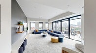 Located at 540 Broadway Street in Saint Paul, MN, this contemporary single-floor amenity space offers a well-appointed layout encompassing a living room, a secondary lounge, a dining area, and a fully equipped kitchen. The interior is defined by clean modern lines, neutral tones, and floor-to-ceiling windows that flood the space with natural light while framing sweeping views of the downtown Saint Paul skyline. The main living area features plush sectional seating, patterned carpet, a wall-mounted TV, and built-in cabinetry, creating a welcoming communal setting. A second lounge area includes a striking stone-surround fireplace with a wood mantel, adding warmth and sophistication. The kitchen is outfitted with flat-front gray cabinetry, white quartz countertops, a subway tile backsplash, stainless steel appliances, and a central island with bar seating. The dining area accommodates a large communal table beneath a sculptural geometric pendant light, complemented by light hardwood flooring. Sliding glass doors open directly onto a spacious rooftop terrace, complete with lounge chairs, outdoor dining tables, a fire pit, and a bar area — all set against a dramatic panoramic view of the Saint Paul cityscape.