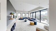 Located at 540 Broadway Street in Saint Paul, MN, this contemporary single-floor amenity space offers a well-appointed layout encompassing a living room, a secondary lounge, a dining area, and a fully equipped kitchen. The interior is defined by clean modern lines, neutral tones, and floor-to-ceiling windows that flood the space with natural light while framing sweeping views of the downtown Saint Paul skyline. The main living area features plush sectional seating, patterned carpet, a wall-mounted TV, and built-in cabinetry, creating a welcoming communal setting. A second lounge area includes a striking stone-surround fireplace with a wood mantel, adding warmth and sophistication. The kitchen is outfitted with flat-front gray cabinetry, white quartz countertops, a subway tile backsplash, stainless steel appliances, and a central island with bar seating. The dining area accommodates a large communal table beneath a sculptural geometric pendant light, complemented by light hardwood flooring. Sliding glass doors open directly onto a spacious rooftop terrace, complete with lounge chairs, outdoor dining tables, a fire pit, and a bar area — all set against a dramatic panoramic view of the Saint Paul cityscape.