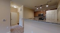 This single-floor residence offers a thoughtfully designed layout that blends functionality with comfort. The kitchen serves as a central highlight, featuring warm honey-toned cabinetry, granite countertops with a subtle speckled pattern, and a full suite of stainless steel appliances including a microwave, electric range, and refrigerator. A breakfast bar extends from the kitchen, providing a seamless transition into the living area while offering additional seating and counter space. Track lighting above the kitchen ensures ample task illumination, complementing the flush-mount ceiling fixtures found throughout the home. Crown molding runs along the ceilings, adding a refined architectural detail that elevates the overall aesthetic. The flooring transitions from plush carpet in the living and entry areas to hard surface in the kitchen zone, offering both warmth and practicality. A hallway off the main living area leads to additional rooms, flanked by tall paneled doors and a built-in storage cabinet that maximizes vertical space. The neutral color palette of warm beige tones on the walls creates a welcoming and versatile backdrop throughout the home. Overall, the space presents a clean, move-in-ready environment with classic finishes and a functional, open-concept feel.