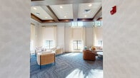 This thoughtfully designed living room offers a refined communal lounge experience, characterized by a striking coffered ceiling with rich dark wood beams, shiplap insets, and recessed lighting that draws the eye upward and enhances the sense of vertical space. Tall windows fitted with sheer drapes and roller shades line the walls, flooding the interior with abundant natural light and offering glimpses of lush outdoor greenery.

The space is divided into several distinct seating arrangements, each with its own character. A central cluster of woven armchairs and sleek accent tables invites casual conversation, while a generous sectional sofa anchors a relaxed lounge area beneath the windows. A dedicated sitting nook features a pair of white accent chairs, a round marble-top coffee table, and a vintage-inspired credenza topped with curated décor and flanked by sculptural gold wall sconces.

A media wall with a flat-screen television is paired with teal-blue cabinetry featuring gold hardware and a white countertop surface, adding a vibrant design accent. The walls are dressed in a soft geometric patterned wallcovering in muted sage and cream tones. Patterned blue-grey carpeting runs throughout, tying the various zones together cohesively. A gallery wall of black-and-white framed photographs adds an artistic, cultural dimension to the overall ambiance.