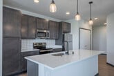 This single-floor, 2-bedroom, 2-bathroom residence offers a clean, modern aesthetic with a practical and comfortable layout. The open-concept living area features light wood-style flooring, a ceiling fan, and a sliding glass door with vertical blinds that opens to an outdoor view, filling the space with natural light. The kitchen is a standout feature, boasting rich dark wood cabinetry, white subway tile backsplash, stainless steel appliances including a refrigerator, range, and microwave, and a large kitchen island with a white quartz countertop illuminated by pendant lighting. In-unit laundry is conveniently situated just off the kitchen. The spacious primary bedroom features plush carpeting, a ceiling fan, and a wide window with outdoor views, complemented by a generous walk-in closet with wire shelving. The second bedroom also features carpeting and ample natural light. Both bathrooms are finished with light wood flooring, dark cabinetry, and white countertops. One bathroom includes a walk-in shower with glass doors, while the other offers a soaking tub and shower combination. The overall ambiance is bright, neutral, and move-in ready, with consistent modern finishes throughout.
