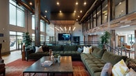 Located at 44 Braves Ave in Lawrenceville, GA, this expansive clubhouse facility showcases an industrial-meets-rustic design aesthetic defined by soaring double-height ceilings, reclaimed wood wall cladding, and structural columns that anchor each zone of the open-plan layout. The main lobby and lounge area features polished tile and hardwood flooring, large floor-to-ceiling windows that flood the space with natural light, and direct access to an outdoor terrace. Seating vignettes are arranged thoughtfully throughout, with plush sectional sofas, wingback chairs, and accent rugs in warm tones creating inviting gathering spots. A built-in shelving wall with dark wood cabinetry adds both function and visual depth. A standout decorative accent wall pairs reclaimed wood with a bold geometric gold-and-grey pattern, lending an artistic focal point to the lounge. The upper mezzanine level, enclosed in steel-framed glass, overlooks the main floor and adds architectural drama. A dedicated game room features a full poker table surrounded by tufted leather seating and a mounted flat-screen TV. The sports lounge area includes an exposed brick accent wall, bar-height seating, multiple large-screen TVs, and a casual lounge corner — ideal for watching live events. Throughout, recessed lighting and pendant fixtures complement the warm, welcoming ambiance.