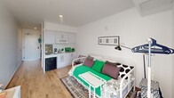 Located at 280 N Oakland Ave in Pasadena, California, this thoughtfully designed studio maximizes its compact footprint through a smart, open single-floor layout. The main living area features light wood flooring, crisp white walls, and recessed lighting that create a clean, bright atmosphere. A wall-mounted adjustable sconce and a mini-split HVAC unit provide functional comfort without sacrificing wall space.

The kitchenette is integrated seamlessly along one wall, featuring white cabinetry, an over-the-range microwave, a compact sink, and a mini refrigerator — offering essential cooking and storage functionality. The living and sleeping area flows naturally from the kitchenette, making efficient use of the open space.

Two hallway areas connect the main space to the full bathroom, which includes a bathtub and shower combination with a curtain enclosure, accented by black corner shelving. A private balcony accessible through a glass door extends the living space outdoors, offering open views of the surrounding Pasadena neighborhood, mature trees, and abundant natural Southern California sunlight — a notable feature that enhances the overall sense of openness in this well-appointed studio.