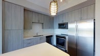 The kitchen in this single-floor space showcases a contemporary design characterized by clean lines and a refined neutral palette. Flat-front cabinetry finished in a warm grey wood-tone lines the walls, providing generous upper and lower storage throughout. The white subway tile backsplash adds a timeless texture that contrasts neatly against the cabinetry, while light-toned countertops extend across the perimeter and onto a central island or peninsula, offering ample prep space.

A full suite of stainless steel appliances includes an electric range, over-the-range microwave, dishwasher, and a full-size refrigerator, equipping the kitchen for functional, everyday use. The undermount sink is positioned along the lower cabinetry run and paired with a modern faucet. A standout design element is the layered gold pendant light suspended from the ceiling, which introduces a warm, decorative focal point and casts an inviting ambient glow. Recessed track lighting complements the pendant, ensuring the workspace is well-lit. The overall aesthetic is polished and cohesive, balancing practicality with modern style.