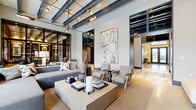This elegantly designed single-floor amenity space offers a refined blend of industrial and contemporary aesthetics across four distinct areas. The entrance corridor sets an immediate tone of sophistication, featuring a striking geometric terrazzo and patterned tile floor alongside sleek black-framed glass partitions that draw the eye toward the main gathering spaces beyond.

The central lounge is a standout, characterized by exposed steel beam ceilings with integrated track lighting, floor-to-ceiling windows that flood the room with natural light, and curated furnishings including plush sofas, textured accent chairs, and a substantial coffee table. Large-scale abstract artwork anchors the walls, while views of an outdoor pool courtyard enhance the open, airy ambiance.

A dedicated billiards room offers a more intimate setting, with a full-size pool table, built-in banquette seating, and oversized wall art framed by linen drapery and natural light from exterior windows.

The bar and social lounge area features polished concrete floors, a sleek bar counter lined with tall white upholstered barstools, dark-toned cabinetry with integrated shelving, wall-mounted televisions, and a cozy dining nook. Adjacent double glass doors provide direct access to a well-equipped fitness center, completing this comprehensive and thoughtfully appointed amenity suite.