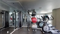 This single-floor fitness space is thoughtfully designed to accommodate a wide range of exercise needs. The room features floor-to-ceiling mirrors along one wall, visually expanding the space while allowing users to monitor their form during workouts. The dark rubber flooring provides a durable, slip-resistant surface suitable for high-impact activities.

On the strength-training side, a multi-station cable machine offers versatile resistance training options. A dedicated rack holds an assortment of exercise balls in various sizes, providing options for core and flexibility training. The cardio section is equipped with at least one elliptical trainer and additional cardiovascular machines, positioned near a window that allows natural light to filter into the space.

Overhead, recessed fluorescent lighting ensures consistent illumination throughout the room, complementing the natural light from the window. A textured ceiling and visible ventilation grilles contribute to a practical, functional aesthetic. Overall, the space is organized efficiently, making it suitable for both individual strength training and cardiovascular exercise within a compact but well-appointed environment.