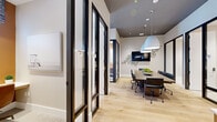 Located at 11590 SW Roma Way in Port Saint Lucie, Florida, this contemporary office space offers a thoughtfully designed single-floor layout built for productivity and collaboration. The central conference area serves as the heart of the space, anchored by a long wood-grain table surrounded by leather task chairs and illuminated by a pair of striking oversized pendant lights. A wall-mounted display screen flanked by decorative botanical dot accents adds both function and visual interest to the meeting area.

Lining the perimeter are several private micro offices, each enclosed by sleek black-framed glass partition walls and doors that maintain an open, airy feel while providing sound separation and focus. Each micro office is outfitted with a built-in desk, task lighting, and curated artwork, creating compact yet fully functional individual workspaces.

Light oak hardwood flooring flows throughout the main areas, contrasting elegantly with the dark ceiling plane and black architectural framing. Warm-toned accent walls within the micro offices add depth and visual contrast. A reception-style credenza with warm wood cabinetry and a clean white countertop provides a practical transition point near the entrance. The overall design strikes a refined balance between open collaboration and private focus, making this a well-appointed professional environment.