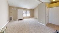 This single-story space presents a thoughtfully designed open-concept floor plan that seamlessly connects the living area and kitchen. The spacious living room is finished with neutral-toned carpet and features vaulted ceilings with exposed beams, lending a sense of openness and volume to the space. A large window with vertical blinds allows natural light to fill the room, enhancing its warm, inviting ambiance. The front entry door is positioned near the window, creating a welcoming point of arrival.

The kitchen is efficiently laid out with dark espresso-toned cabinetry that contrasts cleanly against light countertops and a mosaic tile backsplash in earth tones. White appliances, including a range and refrigerator, are integrated neatly into the design. Wood-look vinyl flooring runs throughout the kitchen, offering durability and visual continuity.

The bathroom is well-appointed with matching dark cabinetry, a vanity with ample counter space, a combined tub and shower enclosed by sliding glass doors, and wood-look flooring that ties the space together. Overall, the unit offers a clean, modern aesthetic with practical finishes throughout.