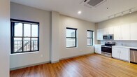 This single-floor unit showcases a clean, contemporary design with a neutral color palette and thoughtful layout. The open living and kitchen area is anchored by warm hardwood-style flooring and multiple large, black-framed windows that frame expansive city skyline views, flooding the space with natural light and providing a strong urban backdrop.

The kitchen is well-appointed with crisp white cabinetry extending to the ceiling, a subway tile backsplash, light countertops, and a full suite of stainless steel appliances, including a refrigerator, electric range, over-the-range microwave, and dishwasher. A ceiling-mounted HVAC unit in the main living area ensures climate control throughout.

The bedroom features plush wall-to-wall carpeting, neutral walls, and a window offering additional city views, creating a calm, comfortable retreat. A second carpeted room provides flexible use as an additional bedroom or den.

The bathroom is finished with large-format light tile flooring, a white vanity with ample cabinet storage, and an accessible grab bar, combining practicality with a clean aesthetic. Overall, the unit offers a cohesive, modern living environment with notable urban views as a defining feature.