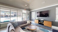 Located at Walkertown Landing in Walkertown, NC, this single-floor community lounge offers a well-appointed, open-concept gathering space designed for both relaxation and productivity. The room features vaulted ceilings with recessed lighting and a sleek ceiling fan, contributing to an airy, spacious feel. Polished concrete floors run throughout, adding an industrial-modern touch that complements the cool gray and white color palette.

The lounge area is anchored by a generous sectional sofa arranged around a round wood coffee table, all centered beneath a wall-mounted flat-screen TV positioned above a warm wood media console. Large floor-to-ceiling glass sliding doors and expansive windows flood the space with natural light and provide direct views of the outdoor pool area, seamlessly connecting the interior to the exterior amenities.

On the opposite end of the space, a built-in workstation features rich wood cabinetry, granite-style countertops, and multiple computer stations, making it a functional resource for residents. A compact kitchenette area with lower cabinetry and a countertop sink rounds out the practical offerings. Decorative framed artwork and wall accents add a polished, welcoming character to this thoughtfully designed communal space.
