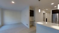 This single-floor space showcases a contemporary open-concept design that seamlessly connects the kitchen and living areas. The kitchen features a white peninsula countertop illuminated by a sleek pendant light, providing both a functional workspace and a natural dividing point between the cooking and living zones. Dark upper cabinetry offers a striking contrast against the bright white walls, adding visual depth to the space.

A distinctive built-in desk nook is integrated into a partial wall, creating a dedicated workspace without occupying additional floor area. This thoughtful architectural detail makes efficient use of the layout while maintaining an open, uncluttered feel throughout.

Light wood-toned flooring extends across the entire space, contributing to a warm and cohesive aesthetic. The walls and ceilings are finished in a clean, bright white, maximizing the sense of openness and allowing natural and artificial light to reflect evenly. Recessed lighting supplements the pendant fixture overhead. A doorway visible in the background suggests additional rooms beyond the main living area, completing this well-designed, functional floor plan.