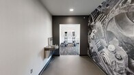This fitness center in Ames, Iowa is a thoughtfully designed, multi-room facility that offers a comprehensive workout experience across three distinct areas. The entry corridor makes an immediate visual impression with a bold, large-scale black-and-white photographic wall mural, textured flooring, and a built-in water fountain — setting an energetic tone from the moment you step inside.

The main workout room is the heart of the facility, finished with patterned commercial carpet in cool gray tones. Large windows line the walls, inviting an abundance of natural light and offering views of the surrounding greenery. This area is equipped with treadmills, elliptical trainers, a multi-station cable machine, a weight bench, and a full rack of dumbbells, providing a well-rounded selection of cardio and strength training options. The neutral gray and white color palette keeps the space feeling clean and focused.

A separate cycling studio offers a change of atmosphere, featuring light hardwood-style flooring, white walls accented with a bold red feature wall, and exposed ductwork that adds an industrial character. Stationary cycling bikes are arranged with ample space for movement.

Together, the rooms create a cohesive, functional, and visually engaging fitness environment suited for a variety of workout routines.