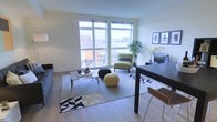 This single-floor space showcases a thoughtfully designed open-plan living and dining area filled with natural light. Expansive floor-to-ceiling windows dominate one wall, offering broad views of the surrounding urban streetscape and flooding the interior with daylight. The light hardwood flooring runs throughout, complementing the neutral wall tones and contributing to a clean, airy atmosphere.

The living area is anchored by a large area rug featuring a bold geometric pattern in black, white, and gold, layered with a glass-topped coffee table and casual pouffe seating in contrasting black and yellow tones. A green accent chair positioned near the windows adds a pop of color alongside a decorative indoor tree, enhancing the fresh, contemporary feel.

Adjacent to the living area, a sleek dark-toned dining table with a barstool provides a defined dining or work zone. A media console positioned against the side wall accommodates a flat-screen television, while framed artwork above adds a curated, modern touch. The overall ambiance is urban, bright, and contemporary, blending functional design with stylish, coordinated décor throughout.