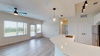This well-designed single-floor residence offers a contemporary open-concept layout that seamlessly connects the kitchen, dining, and living areas. The kitchen is a standout feature, showcasing white shaker-style cabinetry, a light blue subway tile backsplash, stainless steel appliances including a refrigerator, electric range, and microwave, along with a generous curved island topped with a crisp white quartz surface and an undermount sink. Pendant lighting above the island adds a polished finishing touch. Track lighting and recessed fixtures illuminate the space throughout.

Light wood-tone flooring flows through the main living areas, complementing the neutral, warm white walls and creating a bright, airy atmosphere. The living area benefits from abundant natural light through large windows and a set of glass French doors that open to a private balcony with views of the surrounding greenery.

A hallway leads to the bedroom, which is finished with plush carpeting, a ceiling fan, and a window that invites natural light. Adjacent to the bedroom is a spacious walk-in closet with multi-level shelving and hanging rods, offering generous storage capacity. The bathroom features wood-look tile flooring, a tub-shower combination with white subway tile surround, and a wall-mounted storage cabinet.