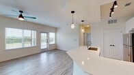 This well-designed single-floor residence offers a contemporary open-concept layout that seamlessly connects the kitchen, dining, and living areas. The kitchen is a standout feature, showcasing white shaker-style cabinetry, a light blue subway tile backsplash, stainless steel appliances including a refrigerator, electric range, and microwave, along with a generous curved island topped with a crisp white quartz surface and an undermount sink. Pendant lighting above the island adds a polished finishing touch. Track lighting and recessed fixtures illuminate the space throughout.

Light wood-tone flooring flows through the main living areas, complementing the neutral, warm white walls and creating a bright, airy atmosphere. The living area benefits from abundant natural light through large windows and a set of glass French doors that open to a private balcony with views of the surrounding greenery.

A hallway leads to the bedroom, which is finished with plush carpeting, a ceiling fan, and a window that invites natural light. Adjacent to the bedroom is a spacious walk-in closet with multi-level shelving and hanging rods, offering generous storage capacity. The bathroom features wood-look tile flooring, a tub-shower combination with white subway tile surround, and a wall-mounted storage cabinet.