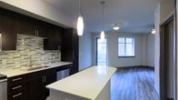 This contemporary single-floor residence showcases a thoughtfully designed open-concept layout that seamlessly connects the kitchen and living areas. The kitchen is a standout feature, boasting rich dark cabinetry with brushed metal hardware, a striking geometric mosaic tile backsplash, and a generous white quartz island that serves as both a prep surface and a casual gathering spot. Stainless steel appliances add a polished, modern touch. Overhead, elegant elongated pendant lights hang above the island, providing focused task lighting while enhancing the overall aesthetic. The flooring transitions smoothly from the kitchen into the living space, where wide-plank hardwood-style floors run throughout, adding warmth and continuity. The living area is bathed in natural light, courtesy of a sliding glass door that opens to a private balcony and an additional window fitted with blinds. The bright, neutral walls and clean architectural lines contribute to a fresh, airy ambiance. The space is well-proportioned, offering a comfortable and functional environment suited to modern everyday living. Overall, the residence delivers a refined balance of style and practicality through its cohesive design choices and quality finishes.