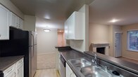 This single-floor residential space offers a practical and comfortable layout designed for everyday living. The kitchen is well-appointed with white cabinetry providing ample storage, dark countertops, a stainless steel double-basin sink, a stainless steel refrigerator, and a built-in dishwasher. The kitchen flows openly into the adjoining living area, creating a connected and convenient floor plan. The living area features a tiled fireplace, adding a warm and inviting focal point to the space. Light-toned flooring runs through the living area, complementing the neutral wall tones throughout. Overhead lighting in the living area provides a soft, even illumination, while the entry door is visible at the far end, contributing to the sense of a well-organized, single-level floor plan. The overall ambiance is clean, understated, and move-in ready, with a cohesive neutral palette that offers a versatile backdrop for a variety of interior styles.