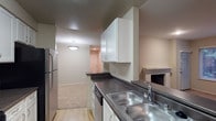 This single-floor residential space offers a practical and comfortable layout designed for everyday living. The kitchen is well-appointed with white cabinetry providing ample storage, dark countertops, a stainless steel double-basin sink, a stainless steel refrigerator, and a built-in dishwasher. The kitchen flows openly into the adjoining living area, creating a connected and convenient floor plan. The living area features a tiled fireplace, adding a warm and inviting focal point to the space. Light-toned flooring runs through the living area, complementing the neutral wall tones throughout. Overhead lighting in the living area provides a soft, even illumination, while the entry door is visible at the far end, contributing to the sense of a well-organized, single-level floor plan. The overall ambiance is clean, understated, and move-in ready, with a cohesive neutral palette that offers a versatile backdrop for a variety of interior styles.