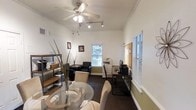 This single-floor office space offers a well-organized and versatile layout designed to support both collaborative and individual work. The main area seamlessly integrates a meeting zone, complete with a round glass-top table and cushioned chairs, alongside a dedicated workstation area furnished with a sleek desk, computer setup, and office equipment including a multifunction printer. A ceiling fan with integrated lighting and a track lighting system ensure consistent illumination throughout, while multiple windows fitted with horizontal blinds allow natural light to filter in and provide views of surrounding greenery. The walls feature a neutral two-tone color scheme, accented by crown molding that adds a subtle architectural detail. An open shelving unit along one wall provides practical storage and display space for office supplies and accessories. A seating area with black upholstered chairs offers an additional space for meetings or brief consultations. A corkboard mounted near the workstation serves as a convenient information hub. The dark carpeted flooring anchors the space with a grounded, professional feel. Overall, the office strikes a balance between functional efficiency and a welcoming, organized atmosphere suitable for a variety of professional uses.