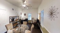 This single-floor office space offers a well-organized and versatile layout designed to support both collaborative and individual work. The main area seamlessly integrates a meeting zone, complete with a round glass-top table and cushioned chairs, alongside a dedicated workstation area furnished with a sleek desk, computer setup, and office equipment including a multifunction printer. A ceiling fan with integrated lighting and a track lighting system ensure consistent illumination throughout, while multiple windows fitted with horizontal blinds allow natural light to filter in and provide views of surrounding greenery. The walls feature a neutral two-tone color scheme, accented by crown molding that adds a subtle architectural detail. An open shelving unit along one wall provides practical storage and display space for office supplies and accessories. A seating area with black upholstered chairs offers an additional space for meetings or brief consultations. A corkboard mounted near the workstation serves as a convenient information hub. The dark carpeted flooring anchors the space with a grounded, professional feel. Overall, the office strikes a balance between functional efficiency and a welcoming, organized atmosphere suitable for a variety of professional uses.