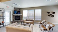 This single-floor space centers around a well-appointed living room that combines comfort with refined design. The focal point of the room is a striking stacked stone fireplace with a crisp white mantel, above which a wall-mounted flat-screen television is prominently positioned. The stone surround adds a natural, textural element that anchors the room with a sense of warmth and character.

Natural light is a defining feature of the living area, courtesy of a set of large windows dressed with horizontal blinds that allow for adjustable brightness and privacy. The neutral taupe wall tones complement the cream and beige upholstered seating, which includes a tufted sofa, matching accent chairs, and sleek modern leather chairs arranged around a rectangular coffee table — creating a balanced, inviting conversation area.

A patterned area rug defines the seating zone and adds subtle visual interest to the light flooring beneath. Recessed lighting in the ceiling supplements the natural light, ensuring the space is well-lit at any time of day. The overall ambiance is cohesive and contemporary, blending transitional design elements to create a living environment that feels both functional and stylish.