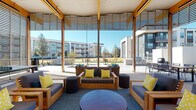 This single-story amenity pavilion is designed to blend indoor comfort with open-air living, featuring a contemporary architectural style defined by exposed wood beams, a white paneled ceiling, and horizontal slatted screens that filter natural light while maintaining expansive views of the surrounding community. Ceiling fans are integrated throughout, ensuring comfortable use across seasons.

The space is organized into distinct zones. An interior lounge area anchors the pavilion with a sleek linear fireplace set into a white paneled wall, wood-framed sofas and armchairs upholstered in deep navy, and accents of teal and yellow through throw pillows and area rugs. A circular coffee table and woven ottomans complete the relaxed seating arrangement.

Opening outward, a semi-covered outdoor lounge continues the wood-framed furniture theme, leading to a generous patio with dining tables, bar-height seating, and built-in grilling stations. Ornamental grasses and decomposed granite landscaping line the walkways, adding a refined, low-maintenance aesthetic.

The exterior of the building features white vertical paneling, wood accents, and black-framed doors and windows, creating a clean and modern curb presence. The overall ambiance is casual yet well-appointed, ideal for communal gathering.