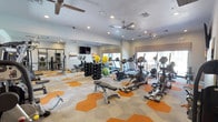 This fitness center offers a comprehensive workout environment designed to accommodate a wide range of exercise routines. The space features a bold, geometric-patterned floor in orange and grey tones that adds a dynamic visual energy to the room. Large windows along one wall flood the interior with natural light, while multiple ceiling fans ensure comfortable airflow throughout workouts.

Full-length mirrors line the walls, enhancing the sense of openness and allowing users to monitor their form during exercise. The cardio section is well-appointed with treadmills, elliptical machines, and a rowing machine, providing ample options for aerobic training. A dedicated free weights area includes a complete rack of dumbbells in varying sizes, accompanied by exercise balls in multiple sizes ideal for strength and stability training.

A weight bench positioned centrally serves as a focal point for resistance training. Recessed lighting supplemented by ceiling fan fixtures ensures the space is consistently well-lit. A wall-mounted television adds entertainment for those engaging in longer cardio sessions. The overall ambiance is modern, clean, and energizing, making this fitness center a fully functional and inviting space for users of all fitness levels.