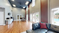 This well-appointed single-floor clubhouse offers a thoughtfully designed open layout that maximizes functionality and comfort. Soaring double-height ceilings create a dramatic sense of openness, accentuated by bold burgundy accent walls and neutral gray tones throughout. Warm hardwood flooring runs continuously across the entire space, tying together each functional zone with visual cohesion.

The main lounge area features a sleek black leather sofa set atop a area rug, accompanied by a wall-mounted flat-screen television, making it an inviting space for relaxation and socializing. Adjacent to the lounge, a kitchenette station is equipped with white cabinetry, a countertop, microwave, coffee maker, and water cooler, providing convenient amenities for guests.

A rustic high-top bar table with bar stools offers additional seating, while French glass doors lead to a separate private room visible in the background. A dedicated fitness area, accessible through another set of glass-panel French doors, is equipped with exercise machines, offering residents a convenient workout space.

Large glass entry doors flood the interior with natural light and open directly onto an outdoor pool courtyard framed by white columns, seamlessly connecting indoor and outdoor living. Recessed and panel lighting throughout ensures bright, comfortable illumination at all times.