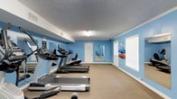 This dedicated fitness center offers a focused and functional workout environment designed to accommodate a variety of exercise routines. The room is finished with bold blue walls that create an energizing atmosphere, complemented by a textured ceiling and neutral carpeted flooring that provides comfort underfoot during workouts.

The cardio area is a central highlight, featuring a lineup of treadmills and additional cardio equipment positioned along one wall, giving users ample space to exercise without feeling cramped. A large mirror is mounted on the wall, allowing users to monitor their form during workouts and also enhancing the sense of openness within the room.

Natural light filters in through a window fitted with white blinds, contributing to a bright and welcoming environment. Along the opposite wall, a yoga mat storage rack keeps the space organized, with rolled mats readily available for floor-based exercises and stretching routines.

Ventilation is supported by ceiling vents, ensuring the space remains comfortable during intense workouts. A private door leads to an adjacent area, adding to the overall functionality of the facility. The layout is clean and practical, making this fitness center a convenient and well-maintained amenity suited for daily use.