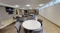 This single-floor community space is thoughtfully designed to accommodate group gatherings, meetings, and social events. The main gathering area is generously proportioned, featuring durable wood-look flooring, a dropped acoustic tile ceiling with recessed and flush-mount lighting, and a row of clerestory windows along one wall that allows natural light to filter into the space. Multiple round white tables paired with upholstered grey chairs are arranged throughout, providing flexible seating configurations to suit a variety of events. Framed artwork and potted plants add warmth to the otherwise neutral, modern interior.

Adjacent to the main room is a dedicated serving area, complete with an extended run of warm wood cabinetry, black granite-style countertops, and a built-in sink — well-suited for buffet-style service or event setup. A pass-through window connects this area directly to the gathering room for convenient access.

A fully equipped kitchen supports the space, outfitted with matching wood cabinetry, black countertops, a full-size refrigerator, a range, and a sink. Durable tile flooring and a wood-paneled door with a glass insert complete the functional, clean aesthetic. Together, these areas create a well-rounded, practical venue suitable for community dining, corporate events, or social programming.