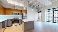 Located on Smith Street in Houston's vibrant Midtown district, this one-bedroom, one-bathroom unit showcases a contemporary industrial aesthetic characterized by soaring ceilings with exposed ductwork, light wood-look flooring, and floor-to-ceiling windows that frame striking city skyline views. The open-concept living and dining area is bathed in natural light, creating an airy, expansive atmosphere despite the unit's efficient footprint.

The kitchen is a standout feature, outfitted with warm wood cabinetry, sleek gray lower cabinets, a white quartz-topped island, under-cabinet LED lighting, and a full suite of stainless steel appliances including a gas range, microwave, and refrigerator. Pendant lighting adds a refined industrial touch above the island and living space.

A well-lit hallway connects the rooms, lined with recessed lighting and clean white walls accented by black hardware. The bedroom offers built-in closet storage and a large window with urban views. The bathroom is finished in a crisp white palette with black-matte fixtures, a tub-shower combination, and wood-look flooring. A dedicated laundry room with full-size washer and dryer completes this thoughtfully designed unit.