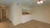 This single-floor residence offers a straightforward and functional layout designed for comfortable everyday living. The main living area is finished with wall-to-wall carpeting in a neutral tone, providing a warm and understated foundation throughout the space. A textured ceiling adds a subtle architectural detail overhead, while a ceiling fan with a light fixture ensures year-round air circulation and ambient lighting.

The kitchen is integrated into the layout via a pass-through counter design, creating an open connection between the cooking and living areas. White cabinetry lines the upper portion of the kitchen nook, offering ample storage, while the countertop extending into the living space provides a convenient surface for dining or casual use. The kitchen is equipped with essential appliances and features a clean, minimal aesthetic consistent with the rest of the space.

Neutral, light-toned walls throughout contribute to a bright and open feel, maximizing the sense of space. HVAC vents are positioned in the ceiling for efficient climate control. A doorway visible from the main living area suggests additional rooms or access points, reinforcing the practical flow of the floor plan. Overall, this residence presents a clean, move-in ready environment with a cohesive and functional design.