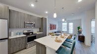 This single-floor residence is designed with a contemporary aesthetic, combining clean lines, neutral tones, and modern finishes throughout. The open-concept kitchen serves as a central feature, showcasing a large island with a white countertop, an undermount sink with a high-arc faucet, and bar seating. Pendant lighting hangs above the island, complementing the recessed ceiling lights. Cabinetry is finished in a warm gray woodgrain tone, paired with a full suite of stainless steel appliances, including a French door refrigerator, electric range, built-in microwave, and dishwasher. Light wood-style flooring flows seamlessly throughout the space.

The living area is bright and open, benefiting from natural light through large windows and a glass-panel door that provides outdoor access. A ceiling fan adds comfort to the space. The bedroom is a well-sized retreat featuring windows with exterior views and ample natural light.

The bathroom is cleanly finished in white, featuring a tub-shower combination, a vanity with dark cabinetry, and wood-style flooring. A doorway leads directly into a walk-in closet equipped with built-in shelving and hanging rods, offering generous storage capacity.
